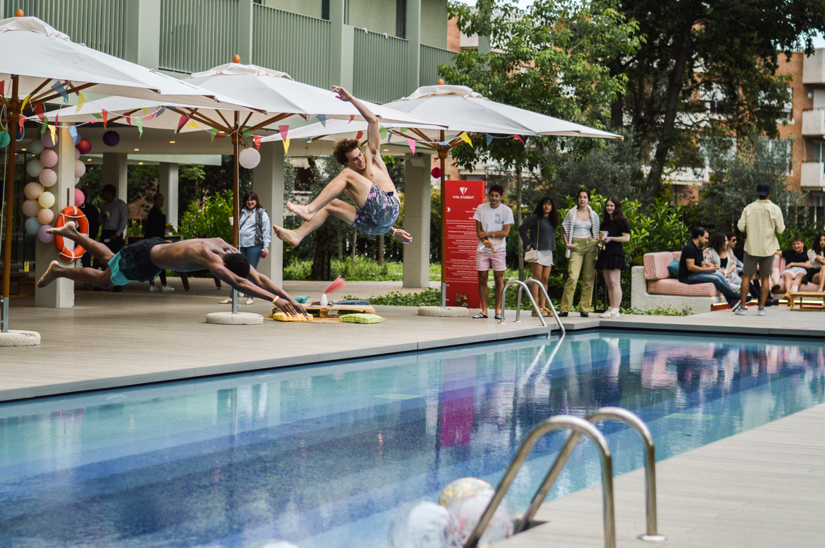 Two young men mid-air, jumping into a swimming pool, surrounded by people socializing. Poolside is decorated with balloons and umbrellas. Some people watch the jump, others sit or stand nearby.